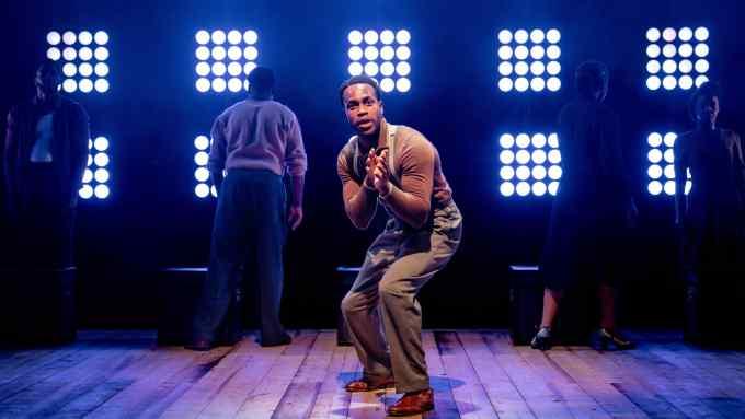 A young man stands on a stage backlit by rectangular clusters of lights; he leans forward eagerly; behind him in shadow, figures have their backs to him