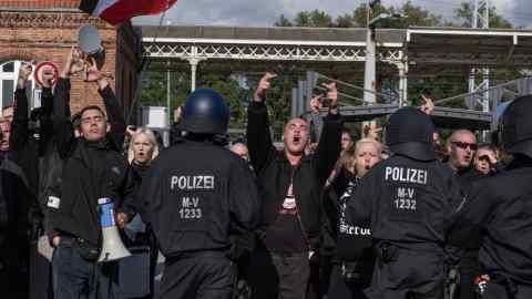 A few police officers, seen from behind, with ‘polizei’ on their backs, restraining a small crowd of demonstrators shouting and making violent hand gestures