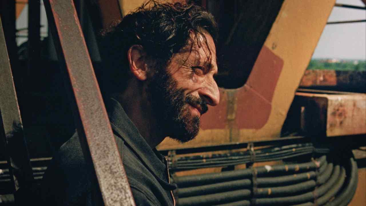 A close-up of a man with dark tousled hair and a face gleaming with sweat and a backdrop of girders and cables